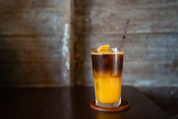 Ein Glas mit Eiskaffee und Mangosaft, Eiswürfeln, Orangenscheibe und Strohhalm steht auf einem Tisch.