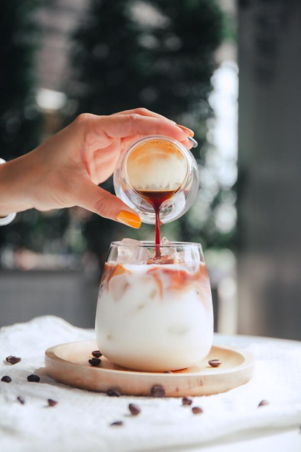 Eine Hand gießt Espresso in ein Glas mit Eiswürfeln und Milch, das auf einem Holzuntersetzer steht.