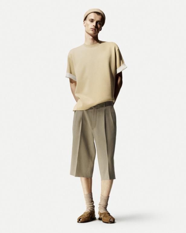 Ein Mann mit Mütze trägt ein beiges T-Shirt, kurze Hosen, Socken und Sandalen.