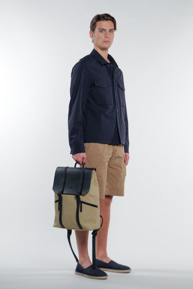 Ein Mann mit marineblauer Jacke und Shorts hält einen beigen Rucksack mit schwarzen Akzenten.