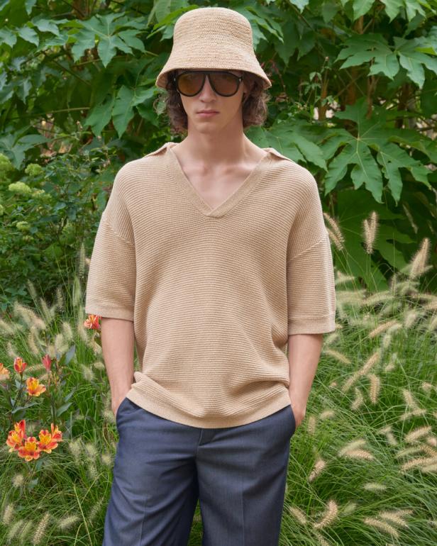 Ein Mann mit beigem Hut, Sonnenbrille und beigem Pullover steht in einem Garten mit grünen Pflanzen und orangefarbenen Blumen.