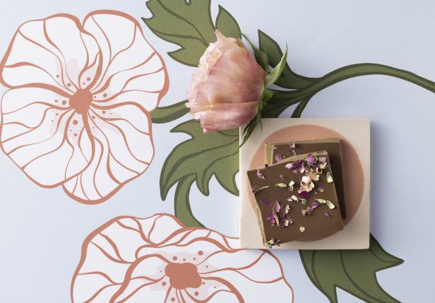 Zwei Schokoladenstücke mit getrockneten Blüten liegen neben einer rosa Rose auf floral gemustertem Untergrund.