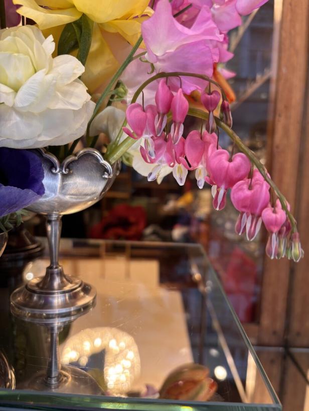 Ein silberner Kelch mit bunten Blumen, darunter auffällige rosa Tränendes Herz-Blüten, steht auf einer Glasfläche.