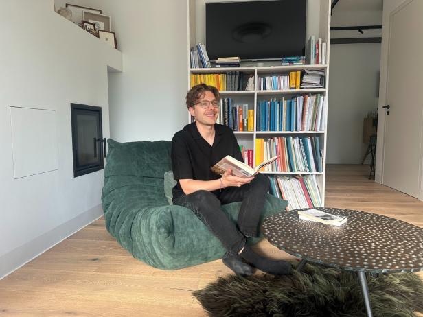 Ein Mann sitzt lesend auf einem grünen Sessel vor einem Bücherregal in modernem Wohnzimmer.