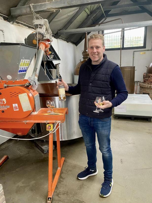 Brandon Carty präsentiert in der Killowen Distillery eine Flasche Whiskey und zwei Gläser vor einer Destillationsanlage.