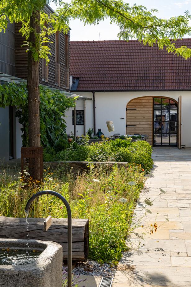 Ein kleiner Innenhof mit Natursteinweg, Brunnen, Sitzbank aus Holz und üppig bepflanztem Garten, umgeben von Gebäuden.