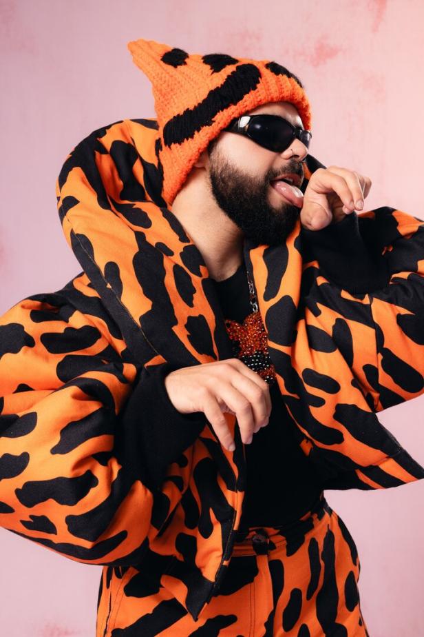 Griechenlands Song-Contest-Teilnehmer Akylas posiert in orange-schwarzem Kostüm und leckt sich über die Hand. 