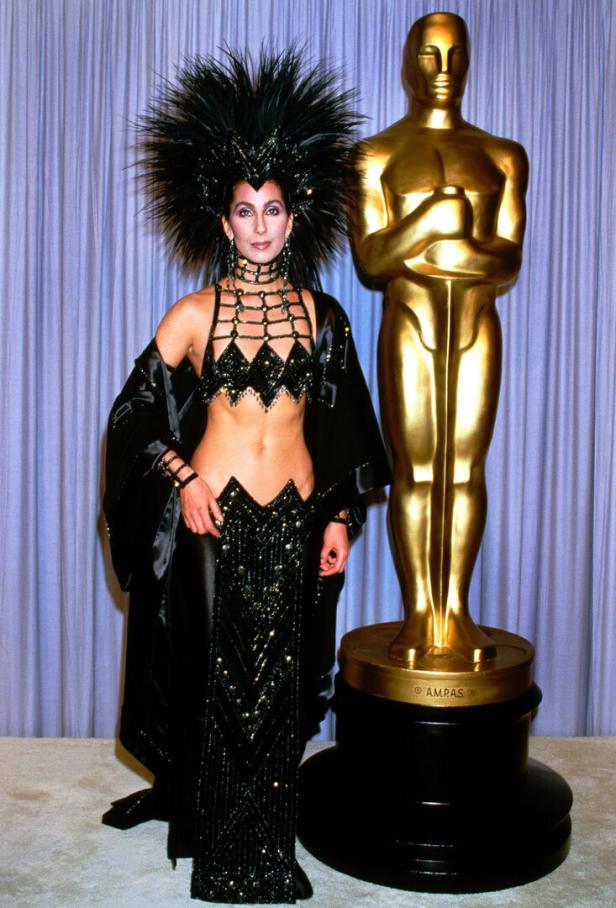 Cher posiert in einem extravaganten, schwarzen Outfit mit Federschmuck neben einer goldenen Oscar-Statue.