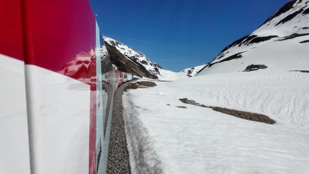 Ein roter Zug fährt durch eine verschneite Berglandschaft unter blauem Himmel.