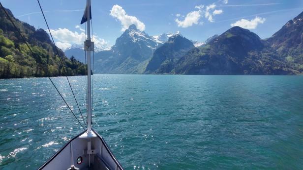 Blick vom Boot auf einen See, dahinter grüne Hügel und schneebedeckte Berge unter blauem Himmel.