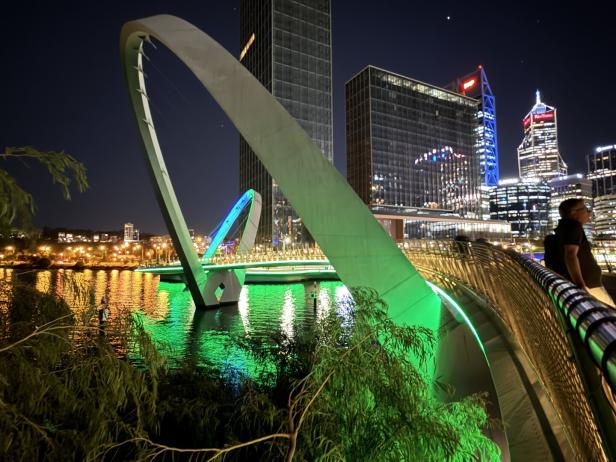 Die erleuchtete Brücke über den Fluss spiegelt sich im Wasser, im Hintergrund die Skyline der Stadt.