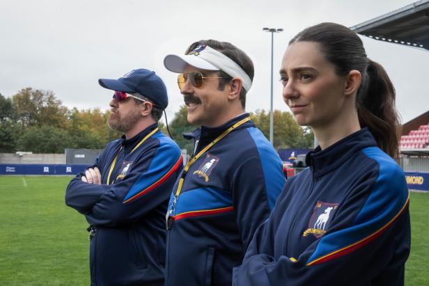 Drei Personen in blauen Trainingsanzügen stehen nebeneinander auf einem Fußballfeld und blicken konzentriert nach vorne.