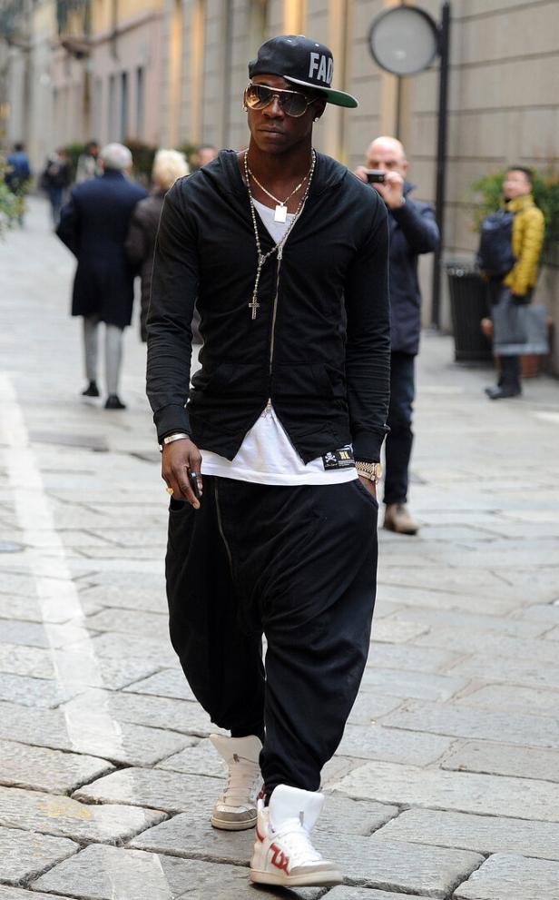 Mario Balotelli mit schwarzer Kappe, Sonnenbrille und auffälliger Kette trägt schwarze Kleidung und weiße Turnschuhe auf einer Straße.