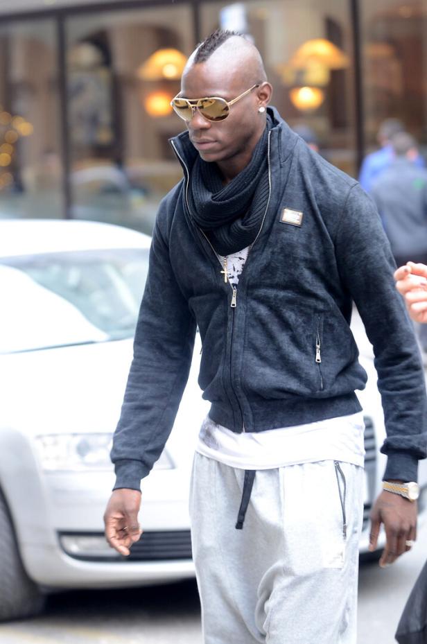 Mario Balotelli mit Sonnenbrille, schwarzer Jacke und grauer Jogginghose geht auf einer Straße entlang.