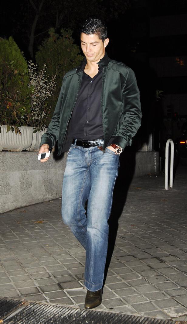 Cristiano Ronaldo mit dunkler Jacke und Jeans geht abends auf einem gepflasterten Gehweg und hält ein Handy in der Hand.