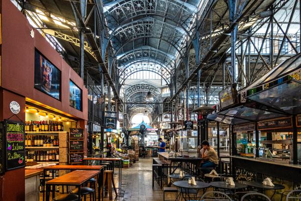 Eine große Markthalle mit hohen Stahlbögen, Verkaufsständen, Regalen voller Waren und wenigen sitzenden Menschen.