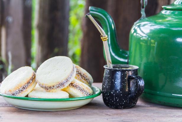Ein Teller mit Alfajores, daneben eine schwarze Tasse mit Metallstrohhalm und eine große grüne Teekanne auf Holztisch.