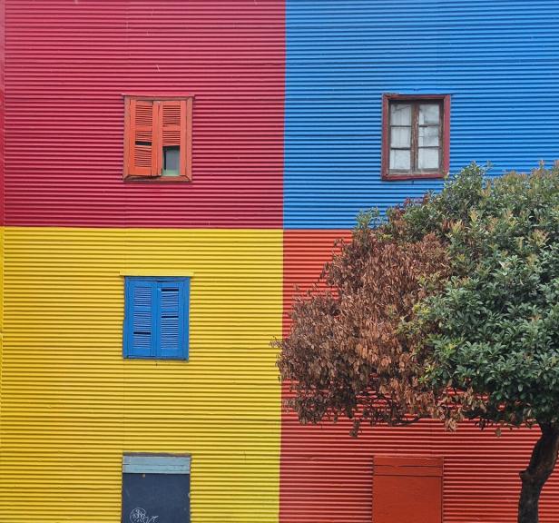 Fassade mit vier farbigen Quadraten in Rot, Blau, Gelb und Orange, mehreren Fenstern und einem Baum rechts.