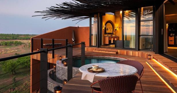 Eine luxuriöse Terrasse mit kleinem Pool, Holzboden, Esstisch und Blick auf eine grüne Savannenlandschaft bei Sonnenuntergang.