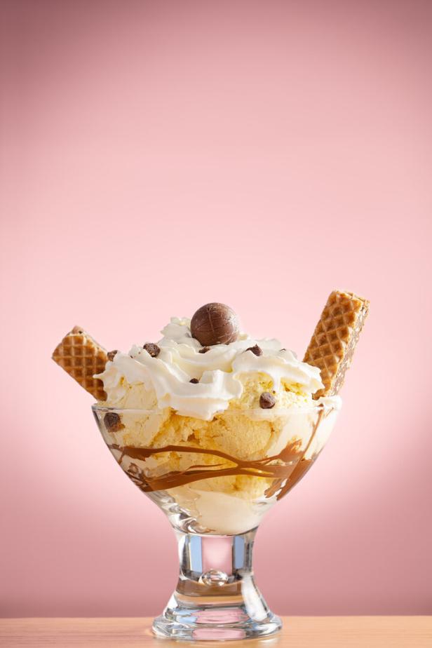 Eisbecher mit Vanilleeis, Schlagobers, Schokostückchen, Waffeln und einer Praline auf rosa Hintergrund.