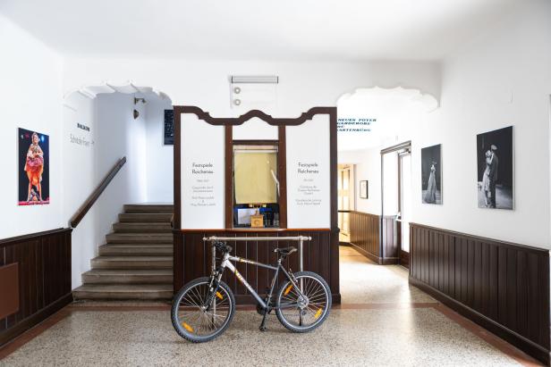 Ein Fahrrad steht vor dem historischen Kassenhäuschen der Festspiele Reichenau in einem hellen Foyer mit dunkler Holzvertäfelung.