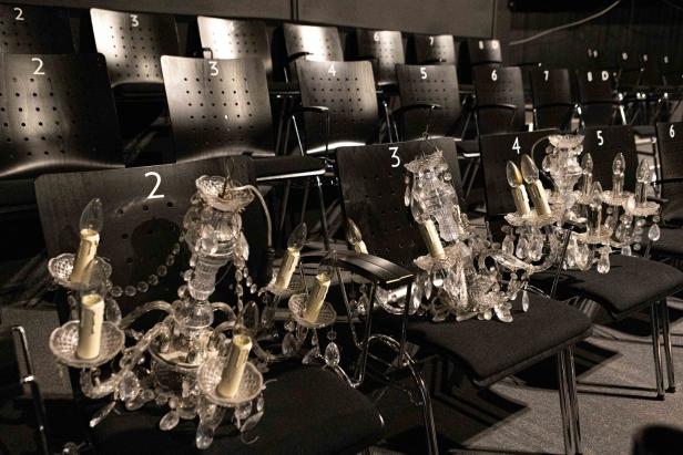 Mehrere verzierte Kristallleuchter stehen auf nummerierten schwarzen Stühlen in einem leeren Theatersaal.
