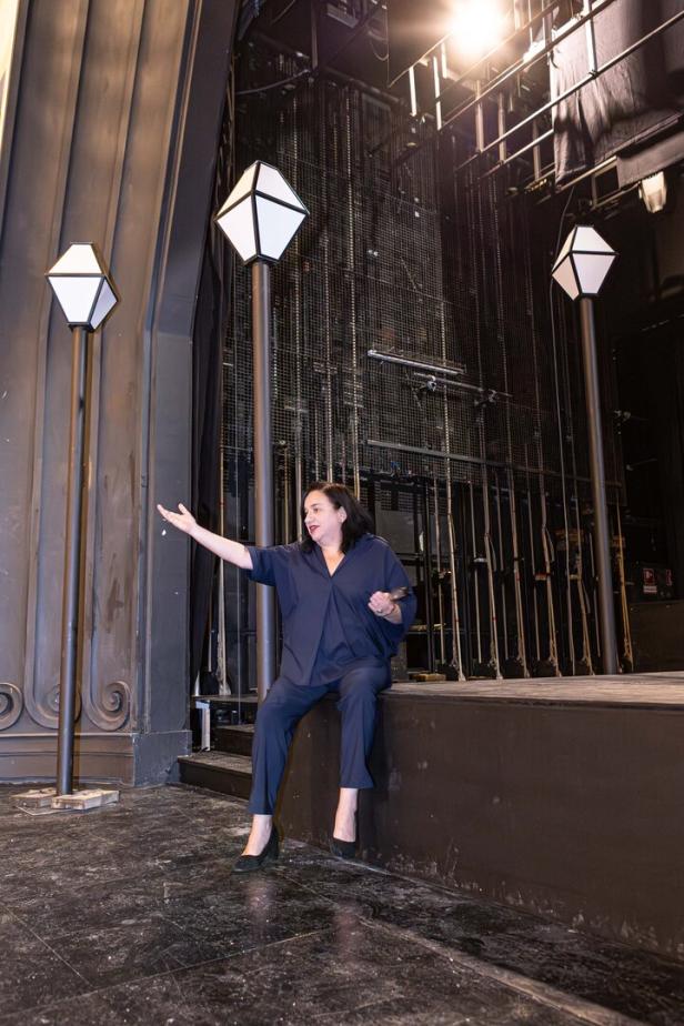 Maria Happel  in dunkler Kleidung sitzt gestikulierend am Bühnenrand eines Theaters, umgeben von hohen Laternen.