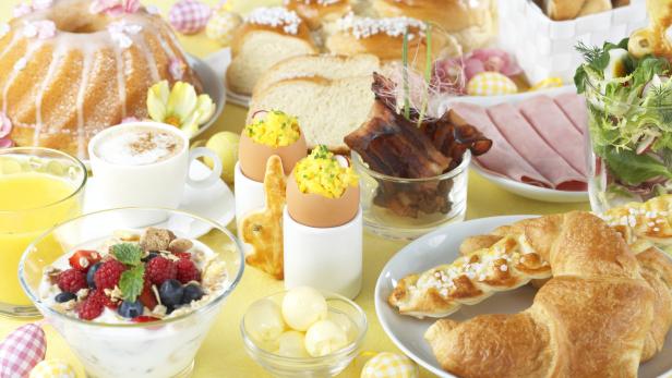 Ein reichhaltiges Osterfrühstück mit Kuchen, Croissants, Eiern, Müsli und Aufschnitt auf einem gelben Tisch.
