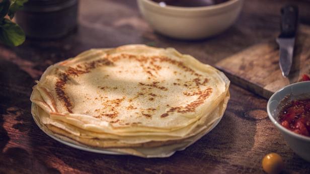 Ein Stapel Palatschinken liegt auf einem Teller, daneben stehen Schalen mit Saucen auf einem Holztisch.