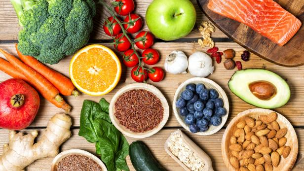 Verschiedene gesunde Lebensmittel wie Gemüse, Obst, Nüsse, Lachs, Reis, Haferflocken und Samen liegen auf einem Holztisch.