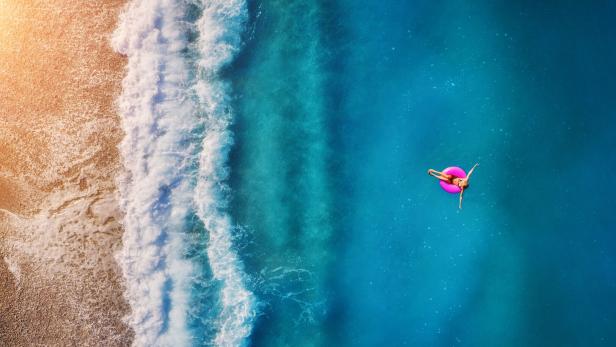Aerial view of young woman swimming on the pink swim ring in the transparent turquoise sea in Oludeniz. Summer seascape with girl, beach, beautiful waves, blue water at sunset. Top view from drone