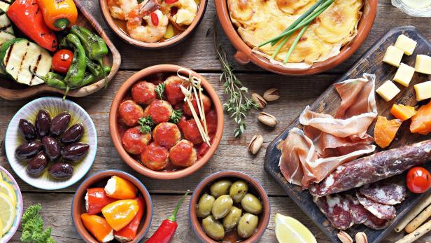 Verschiedene Tapas, darunter Oliven, Fleischbällchen, Käse und Kartoffelgratin, sind auf einem Holztisch angerichtet.