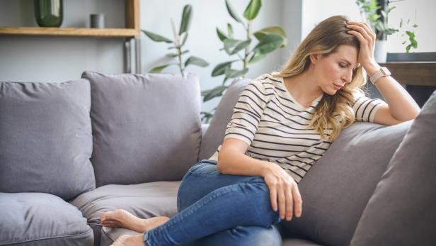 Eine Frau mit blonden Haaren sitzt nachdenklich auf einem grauen Sofa.