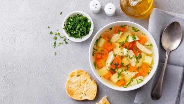 Eine Schale Hühnersuppe mit Gemüse und frischen Kräutern, daneben Brot, Kräuter und ein Löffel auf Serviette.
