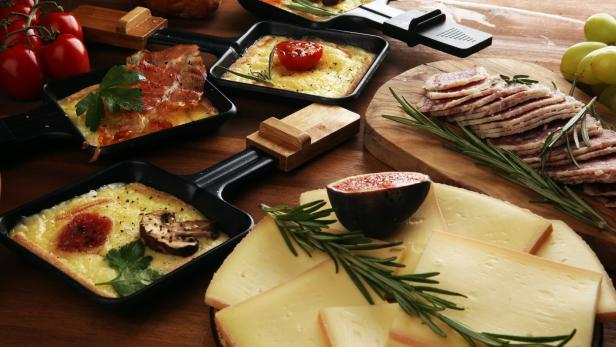 Mehrere Raclettepfännchen mit geschmolzenem Käse, Gemüse und Speck, dazu Käse- und Wurstscheiben mit Rosmarin.