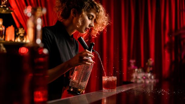 Eine Barkeeperin mixt einen Cocktail vor einem roten Vorhang.