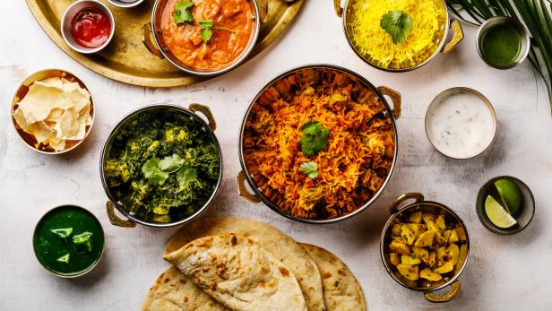 Verschiedene indische Gerichte, darunter Curry, Reis und Brot, sind auf einem Tisch angerichtet.