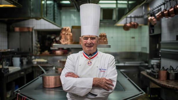 Paul Bocuse, ein Koch mit Kochmütze und Orden, steht in einer Restaurantküche.