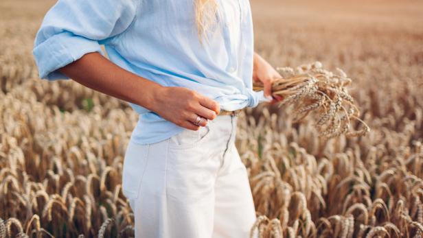 Young woman walking in summer field picking wheat wearing linen shirt and jeans pants. Natural stylish clothes.