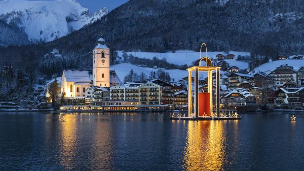 Beleuchtete Kirche und Häuser am verschneiten Seeufer des Wolfgangsees, große schwimmende Weihnachtslaterne auf dem Wasser, Berge im Hintergrund.