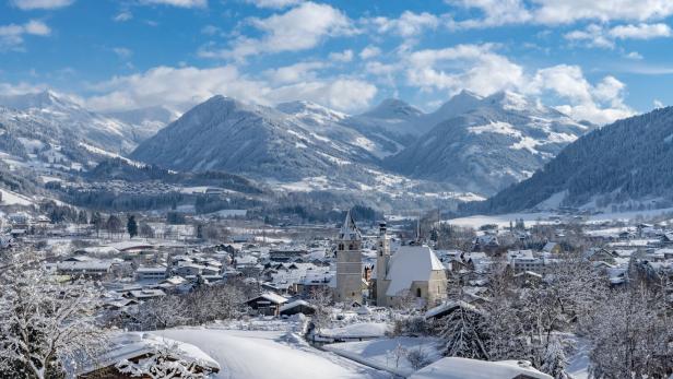 Blick von oben ins Tal auf Kitzbühel: Zwei Kirchen ragen heraus, dahinten erheben sich die Berge. Die Landschaft ist verschneit.