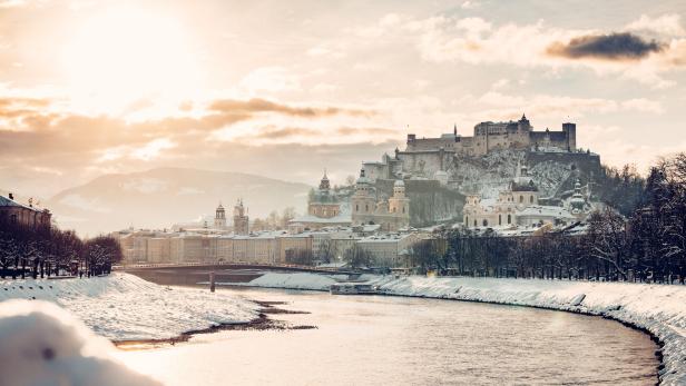Verschneite Stadt mit Fluss, historischen Gebäuden und einer Burg auf einem Hügel im Sonnenlicht.
