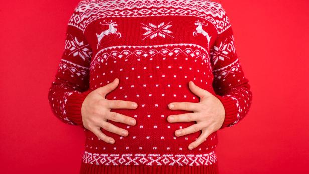 Eine Person im roten Weihnachtspullover hält sich den Bauch vor rotem Hintergrund.