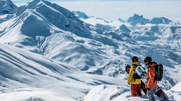 Zwei Snowboarder stehen auf einem verschneiten Gipfel im Skigebiet Arlberg und blicken auf die imposanten Alpen mit langen, leeren Abfahrten.