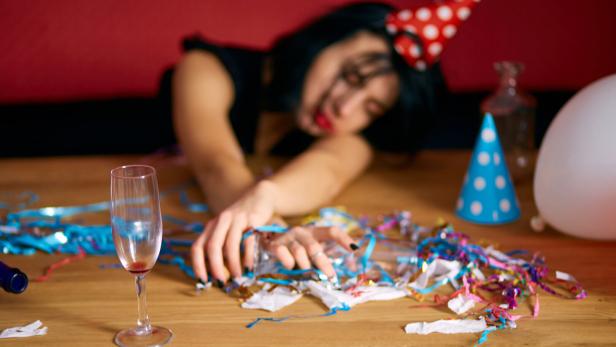 Eine Frau mit Partyhut schläft auf einem Tisch voller Konfetti und Luftschlangen, vor sich ein leeres Sektglas.