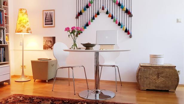 Runder Tisch mit zwei weißen Stühlen, darauf ein Laptop und Blumen, daneben bunte Wanddeko und Bücherregal.