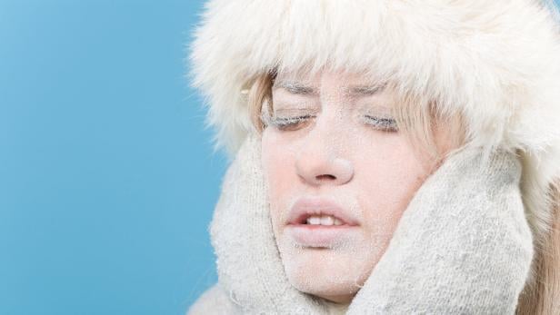Eine Frau mit frostbedecktem Gesicht trägt eine Pelzmütze und Handschuhe vor blauem Hintergrund.