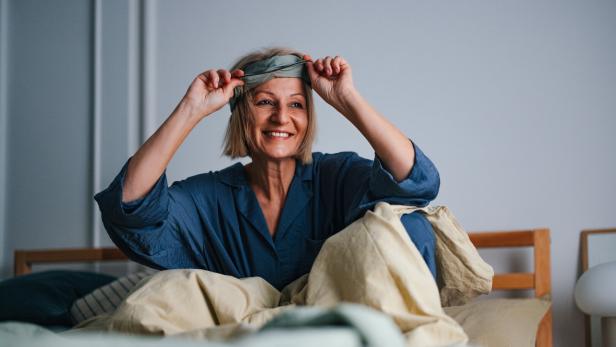Eine lächelnde Frau im blauen Pyjama sitzt im Bett und zieht sich eine Schlafmaske vom Kopf.