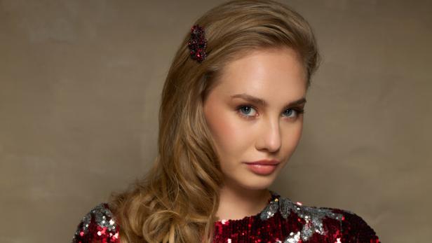 Eine Frau mit blonden Locken trägt ein glitzerndes, rot-silbernes Kleid und eine Haarspange.