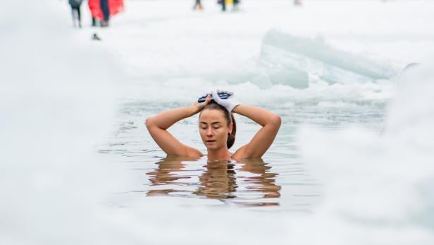 Eine Frau badet in einem eisigen See, umgeben von Schnee, und hält ihre Hände mit Handschuhen auf dem Kopf.